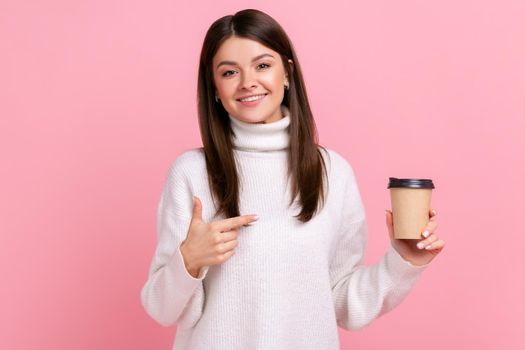 Smiling brunette female holding hot coffee to go, pointing at disposable cup of take away beverage, wearing white casual style sweater. Indoor studio shot isolated on pink background.