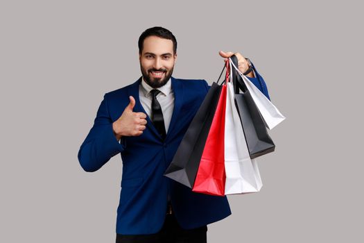 Good looking bearded man showing thumbs up holding shopping bags, pleased with good shopping and discounts, recommending, wearing official style suit. Indoor studio shot isolated on gray background.