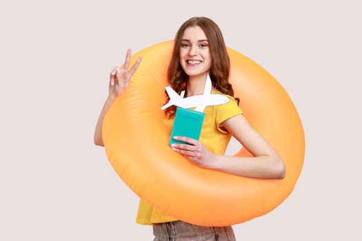 Optimistic tourist teen girl in yellow T-shirt standing with orange rubber ring, holding passport document and airplane mockup, rejoicing travel tour. Indoor studio shot isolated on gray background.