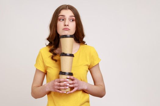 Bored sleepy teenage girl in yellow casual T-shirt holding three coffee, has exhausted expression, bringing drinks in disposable cups. Indoor studio shot isolated on gray background.