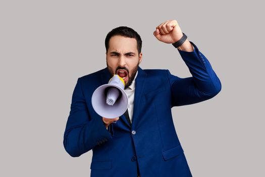 Portrait of bearded businessman standing with raised hands and holding megaphone, screaming in loud speaker, protesting, wearing official style suit. Indoor studio shot isolated on gray background.
