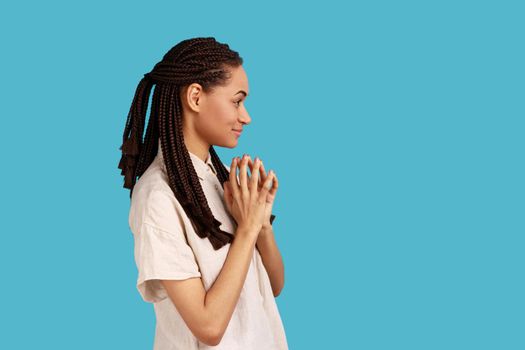 Side view of thoughtful woman with black dreadlocks schemes something, keeps fingers together, considers over cunning plans, wearing white shirt. Indoor studio shot isolated on blue background.