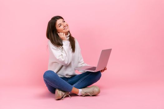 Portrait of female with dreamy facial expression, sitting with crossed legs on floor, holding laptop, wearing white casual style sweater. Indoor studio shot isolated on pink background.