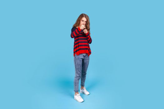 I'll hit you. Full length portrait of aggressive woman standing with raised fists boxing gesture, ready to punch, ready to struggle, fighting spirit. Indoor studio shot isolated on blue background.