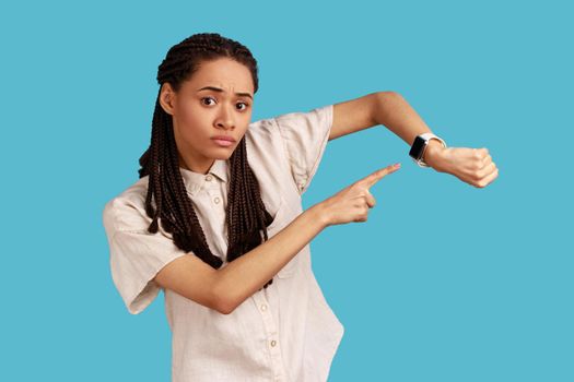 Concerned punctual woman with black dreadlocks pointing finger at smartwatch on her wrist, look at time, hurry up and act, wearing white shirt. Indoor studio shot isolated on blue background.