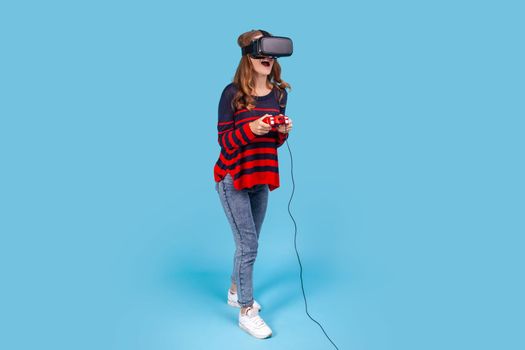 Full length portrait of woman wearing striped casual sweater, VR glasses headset, holds joystick in hands, playing game experiencing, excited expression. Indoor studio shot isolated on blue background