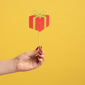 Closeup side view portrait of woman hand holding festive party props, red little paper present box on stick, celebrating holiday. Indoor studio shot isolated on yellow background.