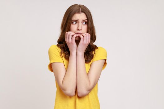 Anxious nervous female of young age with brown hair in T-shirt biting nails on fingers looking aside with terrified expression, confused and worried. Indoor studio shot isolated on gray background.