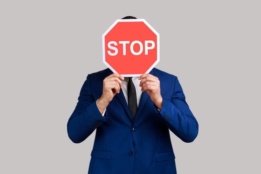 Portrait of man hiding face behind Stop symbol, red traffic sign warning of restricted access, banned service, wearing official style suit. Indoor studio shot isolated on gray background.