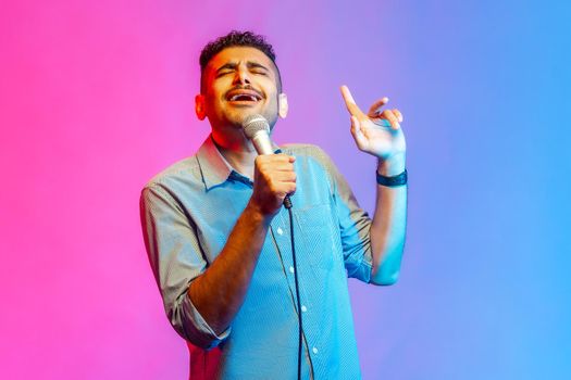 Portrait of happy man in shirt loudly singing song holding microphone in hand, having fun resting in karaoke, singer performance. Indoor studio shot isolated on colorful neon light background.