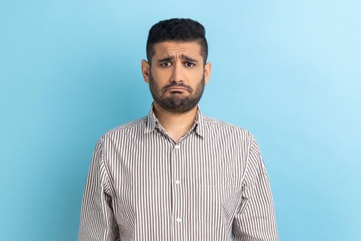 Bearded upset businessman standing and looking at camera with dissatisfied sadness face, expressing sorrow, having bad mood, wearing striped shirt. Indoor studio shot isolated on blue background.