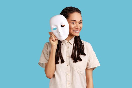 Woman with dreadlocks removing white mask from face showing his smiling expression, good mood, pretending to be another person, wearing white shirt. Indoor studio shot isolated on blue background.