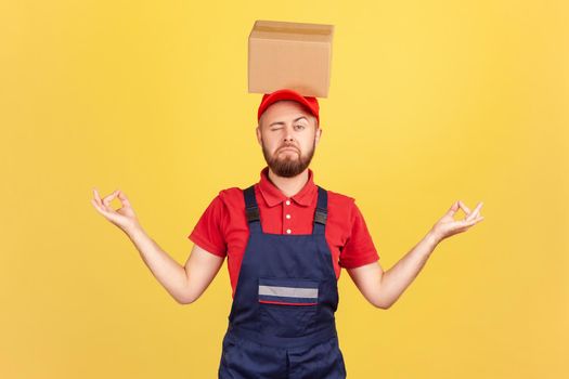 Portrait of relaxed courier man standing with cardboard box on his head, practicing yoga, looking at camera with one eye, calms down. Indoor studio shot isolated on yellow background.