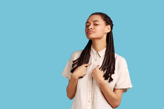 Its me. Boastful woman with dreadlocks pointing at herself, proud of own achievements, looks confidently and self assured at camera, wearing white shirt. Indoor studio shot isolated on blue background