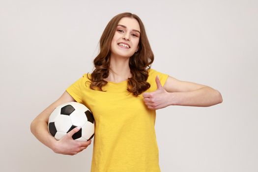 Young beautiful brown haired teenager girl holding soccer ball, looking at camera with toothy smile. showing thumb up, wearing casual attire. Indoor studio shot isolated on gray background.