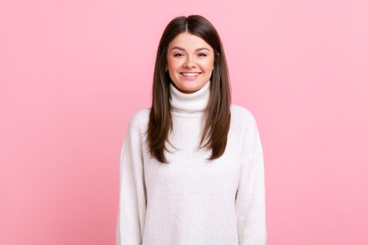 Attractive female with dark hair smiling to camera, expressing positive emotions, being in good mood, wearing white casual style sweater. Indoor studio shot isolated on pink background.
