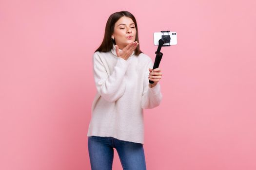 Happy romantic pretty female blogger using smartphone and steadicam for streaming, sends air kisses, wearing white casual style sweater. Indoor studio shot isolated on pink background.