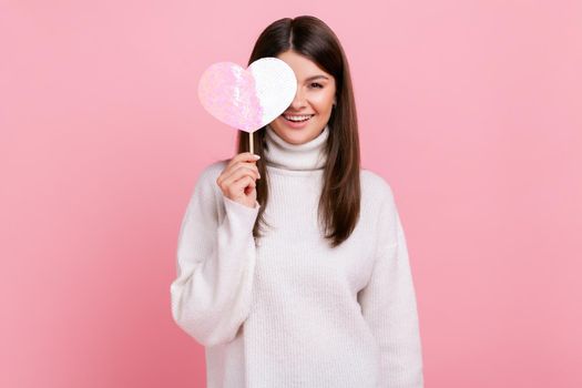 Romantic brunette female standing covering eye with rosy heart on stick, expressing love and romance, wearing white casual style sweater. Indoor studio shot isolated on pink background.