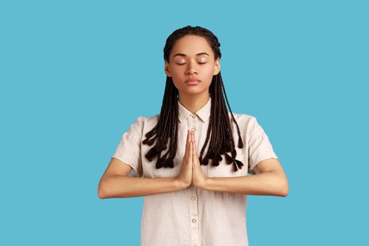 Gorgeous lovely woman with black dreadlocks keeps hands in yoga gesture, has calm facial expression, keeping palms pressed together and closed eyes. Indoor studio shot isolated on blue background.