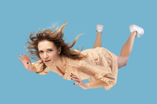 Beautiful young woman levitating in mid-air, falling down and her hair messed up soaring from wind, model flying hovering with dreamy peaceful expression. indoor shot isolated on blue background