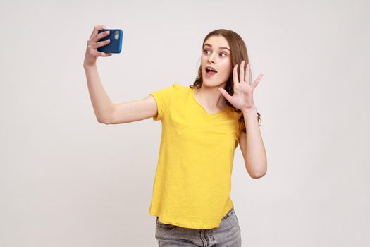 Portrait of happy curly-haired teen girl in yellow T-shirt taking selfie, having conversation on video call and waving hello gesture, vlog broadcast. Indoor studio shot isolated on gray background.