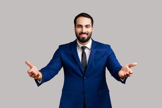 Portrait of smiling satisfied bearded businessman widely spreading his arms welcoming dear guests, greeting, wearing official style suit. Indoor studio shot isolated on gray background.