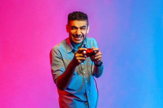 Portrait of amazed man in shirt holding in hands red gamepad joystick, looking at camera with optimistic expression, enjoying game. Indoor studio shot isolated on colorful neon light background.