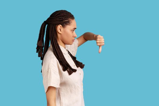 Side view of discontent woman with black dreadlocks showing disapproval sign, keeps thumb down, expresses dislike, frowns face in discontent. Indoor studio shot isolated on blue background.