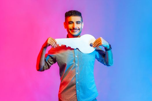 Portrait of positive man in shirt holding huge paper key and smiling satisfied with new home, real estate purchase, rental service. Indoor studio shot isolated on colorful neon light background.