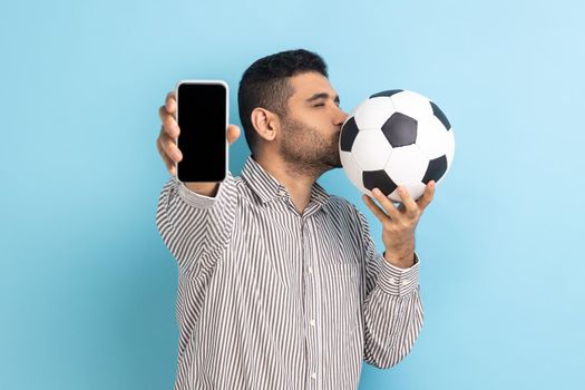 Portrait of bearded handsome man standing kissing soccer ball and showing mobile phone with empty black display, wearing striped shirt. Indoor studio shot isolated on blue background.