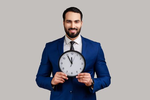 Handsome bearded businessman holding big clock and looking at camera with toothy smile, playful optimistic expression, wearing official style suit. Indoor studio shot isolated on gray background.