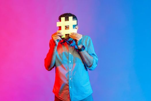 Portrait of young adult man in shirt looking through big hashtag symbol, tagged message, popular internet idea, viral content. Indoor studio shot isolated on colorful neon light background.