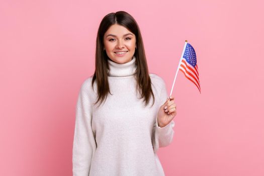 Attractive brunette woman holding American flag in her hand and looking at camera with positive, wearing white casual style sweater. Indoor studio shot isolated on pink background.