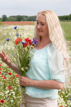 a beautiful middle-aged blonde woman stands among a flowering field of poppy, daisies, Cornflowers, and holds a bouquet of wild flowers and laughs. High quality photo