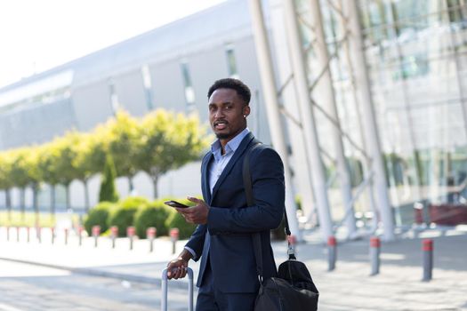 african american business man standing on the background a modern train station airport in formal suit with a suitcase using app mobile phone. Traveling businessman tourist with a smartphone in hands