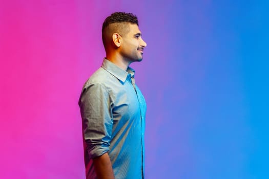Side view portrait of young adult smiling handsome man in shirt standing looking ahead with happy positive facial expression. Indoor studio shot isolated on colorful neon light background.