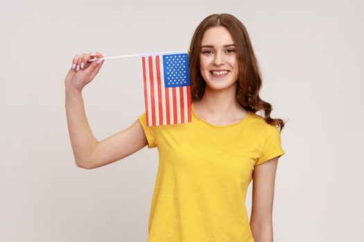 Smiling attractive teenager girl in yellow casual T-shirt holding usa flag looking at camera with toothy smile, human rights, freedom of speech. Indoor studio shot isolated on gray background.