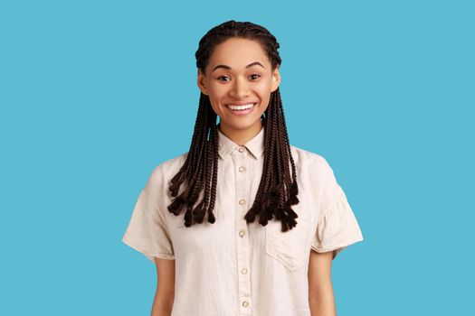 Portrait of cheerful beautiful woman with black dreadlocks, having charming engaging smile and positive emotions, wearing white shirt. Indoor studio shot isolated on blue background.