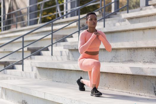 African American woman performs fitness exercises for weight loss, performs squats in a pink suit in the morning in the air near the stadium