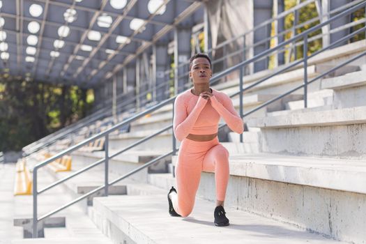 African American woman performs fitness exercises for weight loss, performs squats in a pink suit in the morning in the air near the stadium