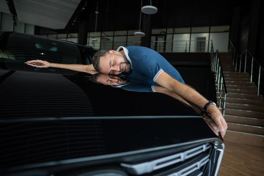 Happy caucasian man hugging the hood of his new car in a car dealership