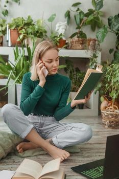 A blonde woman in a room with a lot of green indoor plants is working on a laptop. The concept of biophysical design in the interior. Work from home, work as a freelancer.