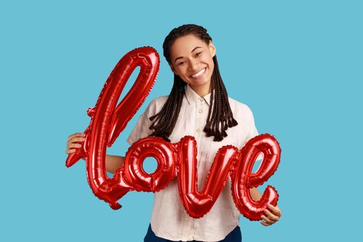 Satisfied attractive woman with black dreadlocks holding love word of foil balloons, expressing romantic emotions and feeling, wearing white shirt. Indoor studio shot isolated on blue background.