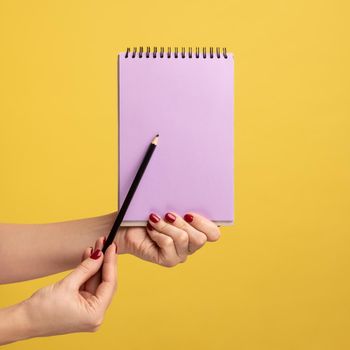 Profile side view closeup of woman hand holding purple notepad in hand and showing empty paper with pen or pencil. Indoor studio shot isolated on yellow background.