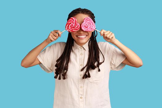 Portrait of playful childish romantic woman with black dreadlocks covering eyes with sugary hear shape candies, wearing white shirt. Indoor studio shot isolated on blue background.