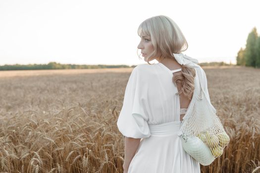 A blonde woman in a long white dress walks in a wheat field. The concept of a wedding and walking in nature.