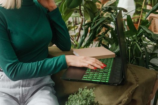 A blonde woman in a room with a lot of green indoor plants is working on a laptop. The concept of biophysical design in the interior. Work from home, work as a freelancer.