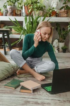 A blonde woman in a room with a lot of green indoor plants is working on a laptop. The concept of biophysical design in the interior. Work from home, work as a freelancer.