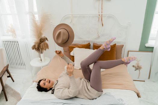 Lifestyle concept. A young middle-aged woman in a sweater lies on the bed and laughs, tossing her brown hat in her hands