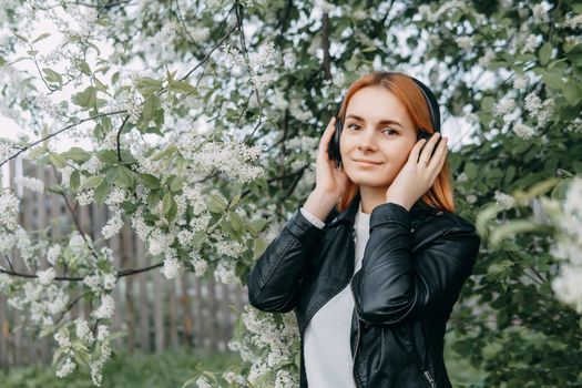 A red-haired teenage girl listens to music in headphones, configured via the phone. The time of spring flowering of cherry orchards.